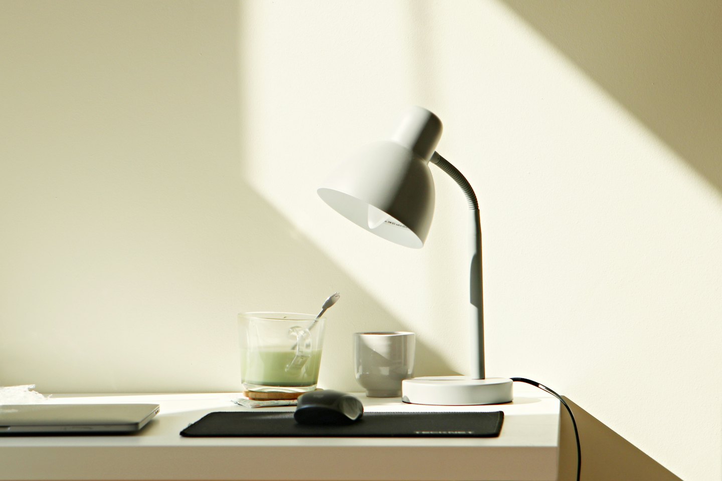 Scrivania con laptop, mouse, lampada e bevanda illuminata dal sole.